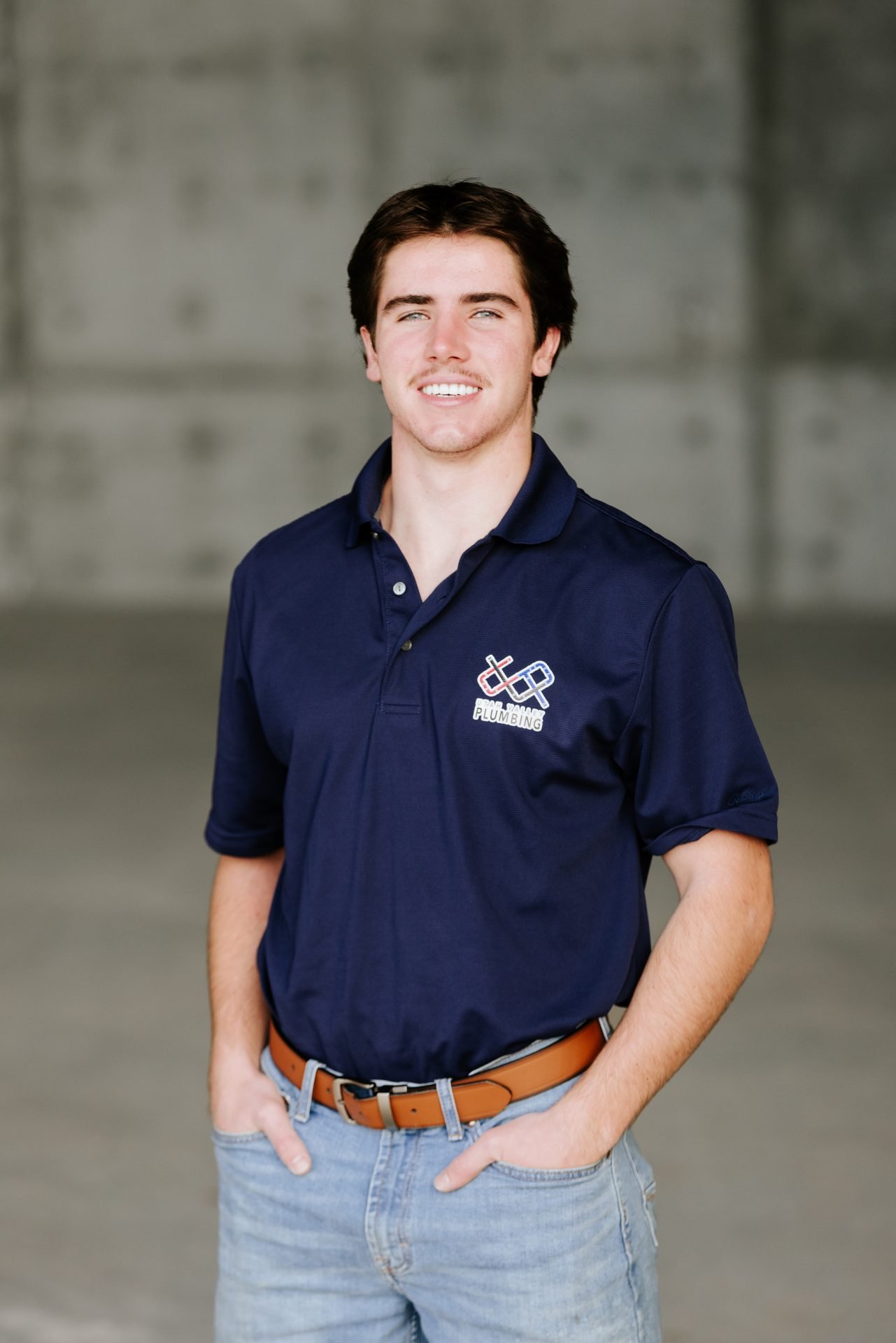 Young man in navy polo shirt with "Plumbing" logo, smiling confidently in a professional setting, representing Utah Valley Plumbing team.