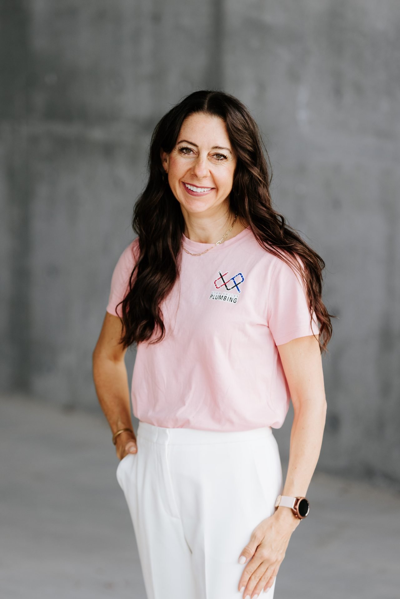 Woman smiling in a pink shirt with a plumbing logo, standing against a gray background, representing Utah Valley Plumbing's team.