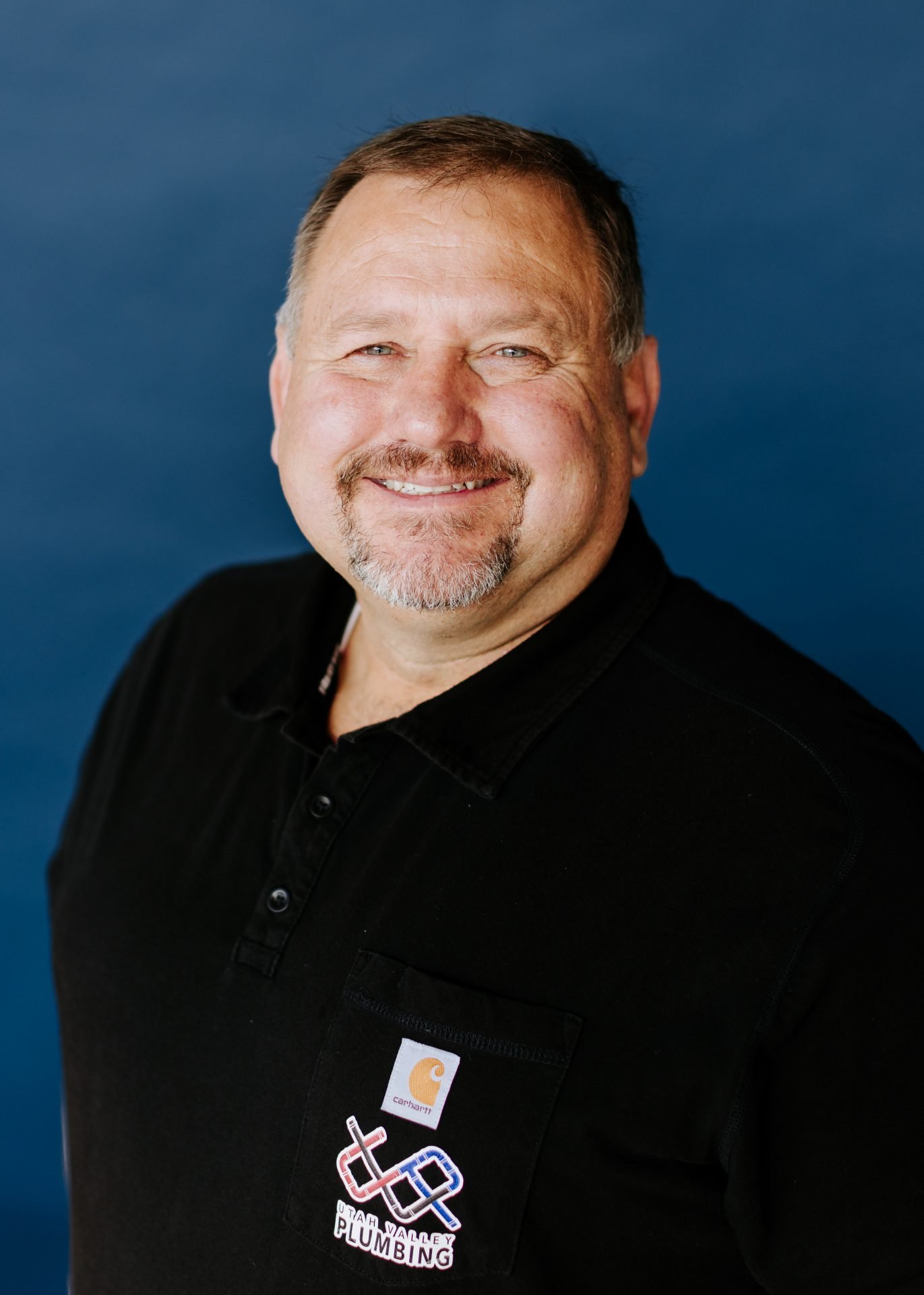 Smiling man wearing a black shirt with "Utah Valley Plumbing" logo, against a blue background, representing the plumbing team.