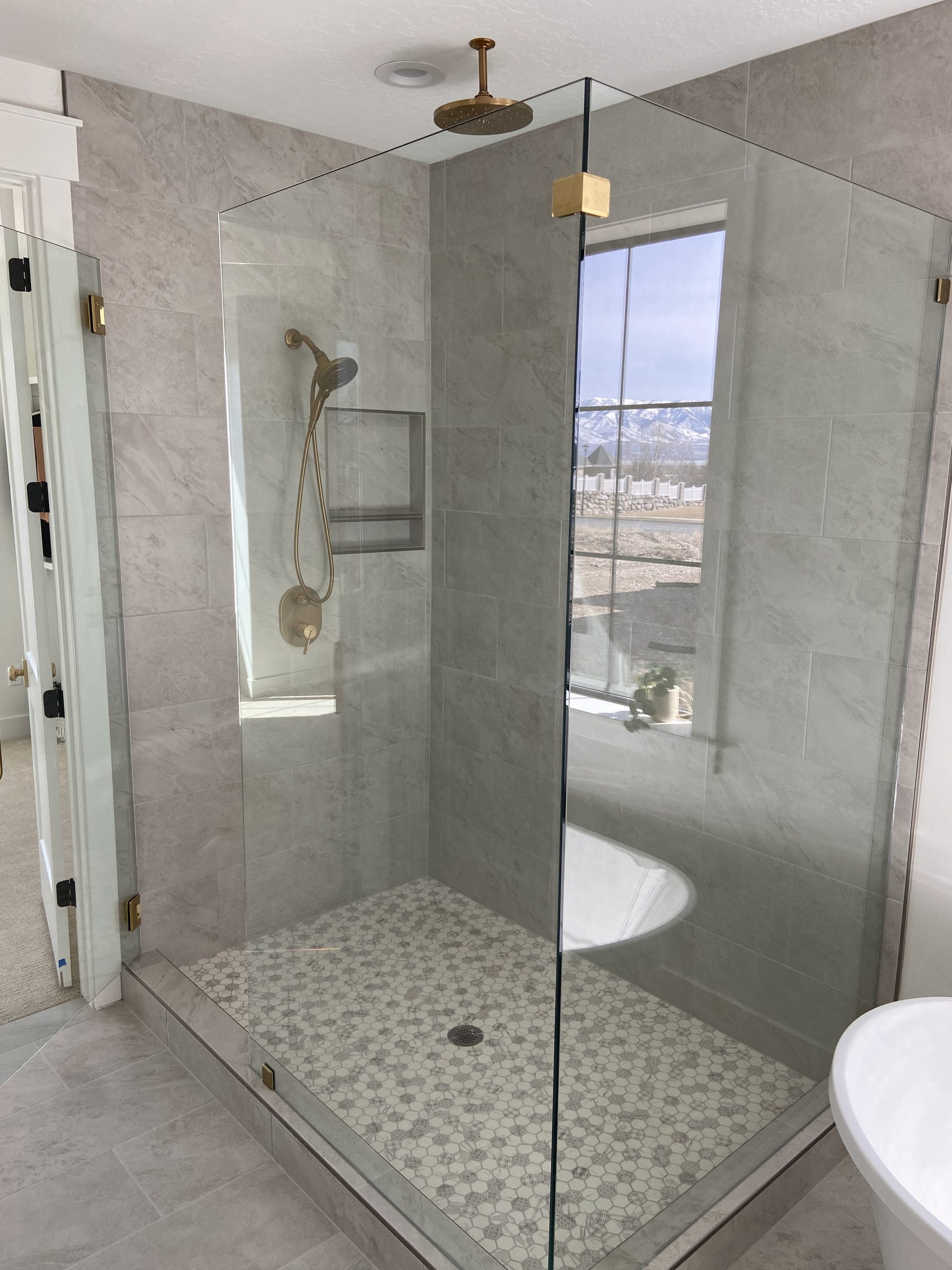 Modern glass shower enclosure with gold fixtures, hexagonal tile floor, and large window showcasing mountain view, reflecting Utah Valley Plumbing's expertise in bathroom renovations.