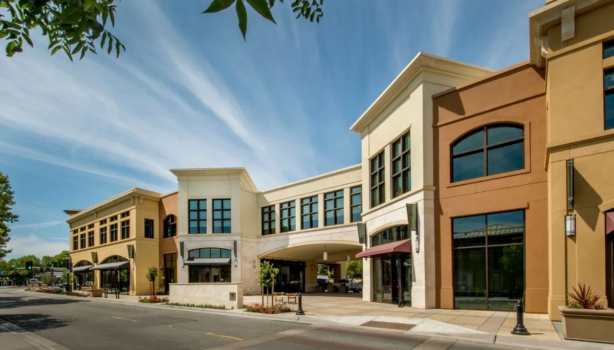 Modern commercial building with large windows, beige and brown exteriors, and an arched driveway entrance under a clear sky.