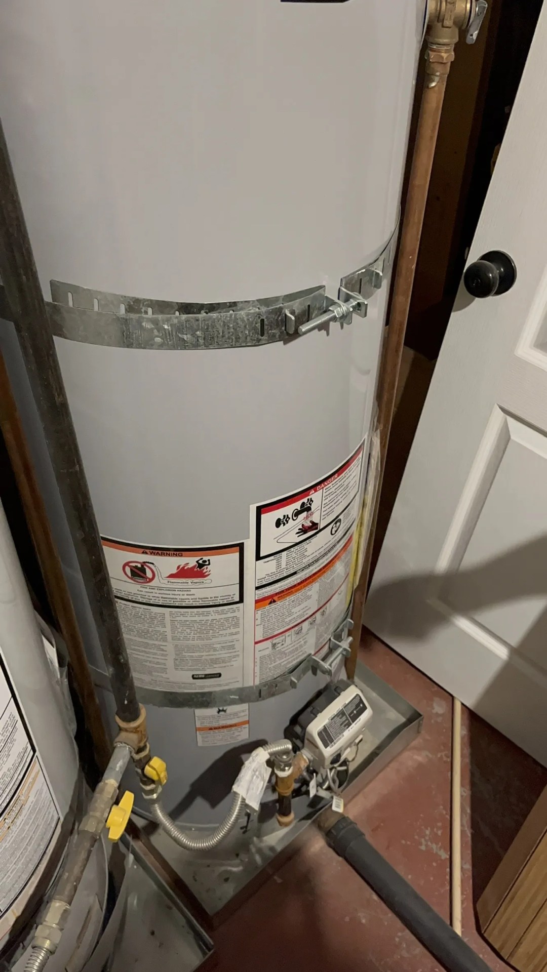 Water heater with safety labels and plumbing connections in a basement setting.